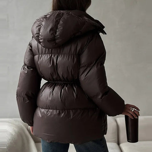 A woman viewed from behind wearing a dark brown puffer jacket with a belted waist and hood, standing indoors near a white sofa and holding a dark tumbler. The brand name 'hfwomenf' appears in the top left corner.