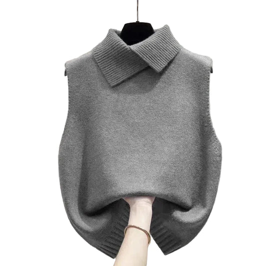 A gray knit sweater vest with a wide ribbed collar, hanging on a black hanger against a beige wall. A hand is holding the hem of the vest to show its drape.