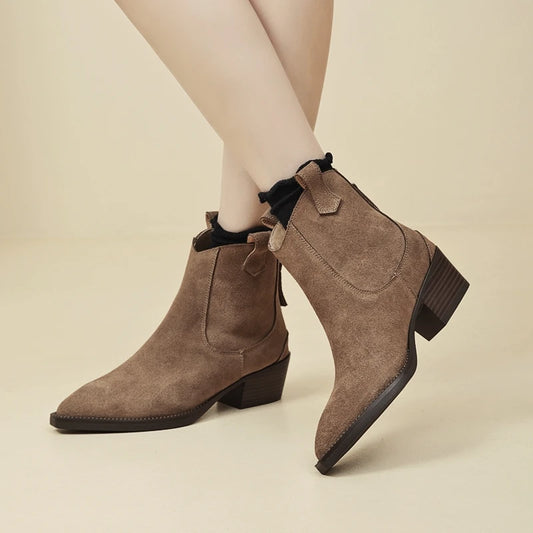 A pair of women's taupe-brown suede ankle boots with black ruffled trim at the collar, shown on a model's feet against a warm beige studio background.