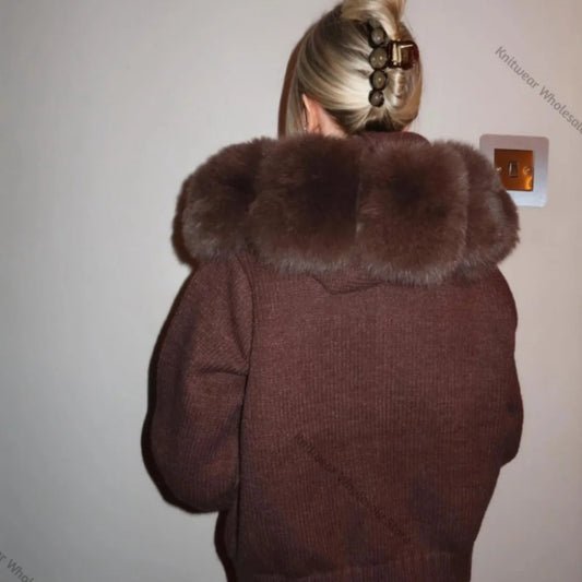 A person viewed from behind wearing a brown knit hooded jacket with a large dark brown faux-fur trimmed hood, standing against a plain light wall.