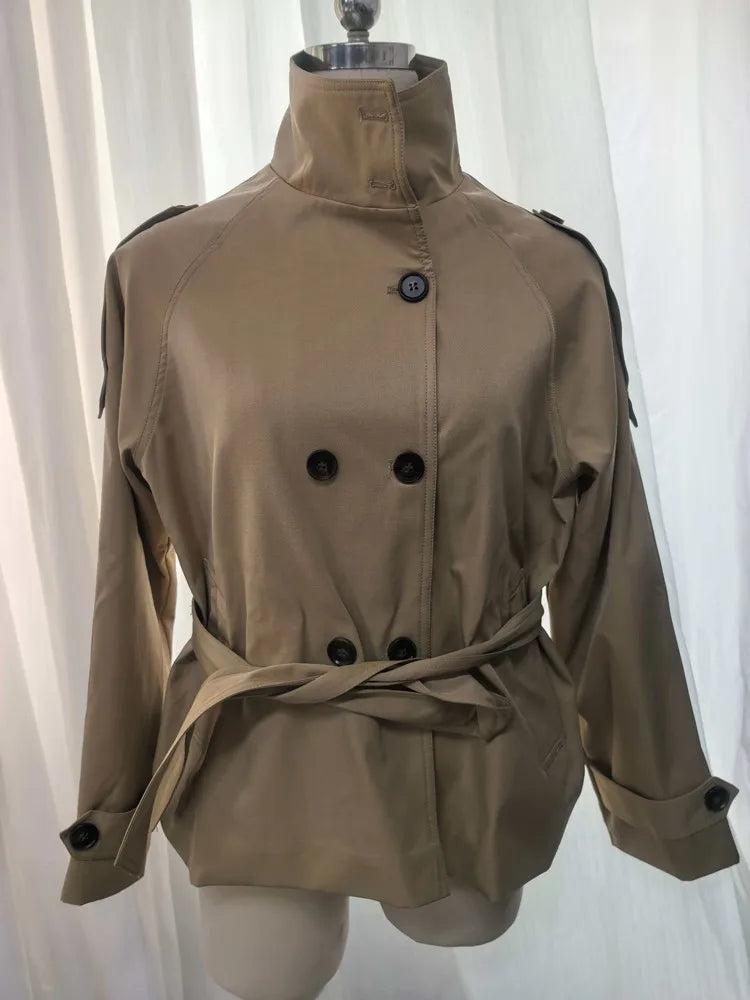 A tan trench coat with a high stand-up collar, double-breasted buttons, shoulder epaulets, and a matching waist tie, displayed on a dress form against a white curtain background.