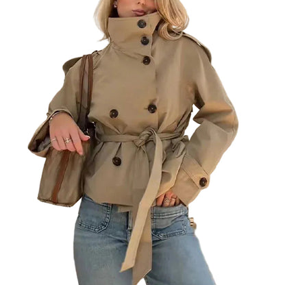 A woman modeling a cropped beige trench coat with a high collar, double-breasted buttons, and a matching waist tie, paired with blue jeans and a brown shoulder bag against a plain white background.