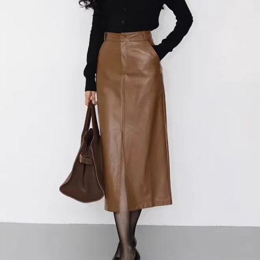 A woman models a high-waisted brown faux-leather midi skirt paired with a black long-sleeve button-up top, black sheer tights, black pointed-toe heels, and a brown leather tote bag against a plain white wall.
