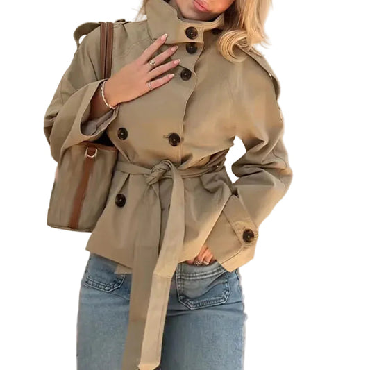A woman wearing a beige double-breasted belted trench coat with dark buttons, paired with blue jeans and carrying a brown shoulder bag, set against a plain white background.