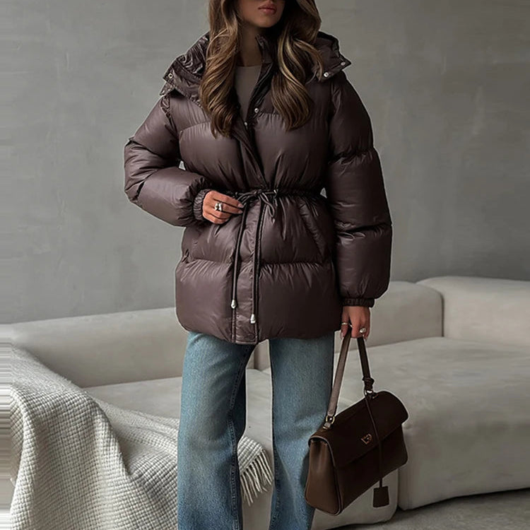 A woman wearing a dark brown puffer jacket with a drawstring waist, blue jeans, and sunglasses, holding a brown handbag in a modern living room setting.