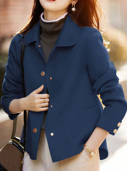 A woman wearing a navy blue tailored jacket with gold-tone buttons over a dark gray turtleneck sweater and light beige pants, photographed outdoors with warm sunlight and a blurred background.