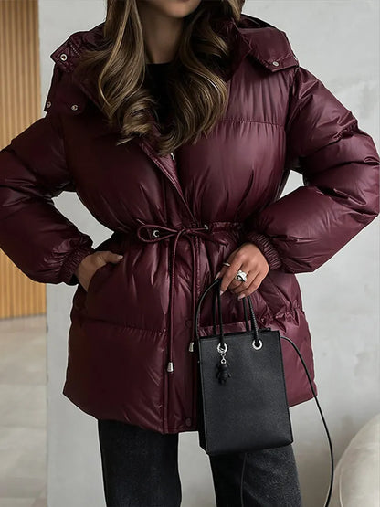 A woman wearing a shiny, quilted puffer jacket in deep burgundy with a drawstring waist, holding a black handbag.