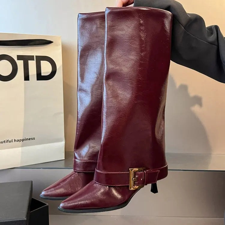 Brown leather knee-high boot with a gold buckle held by a hand, with a blurred background.