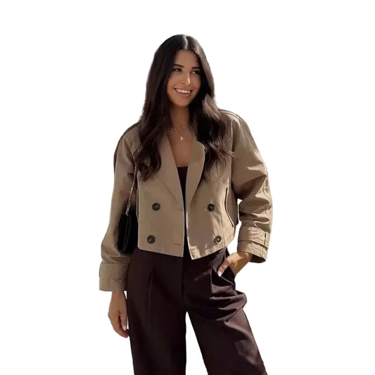 Woman in a beige jacket standing in front of a scenic view with buildings and greenery.