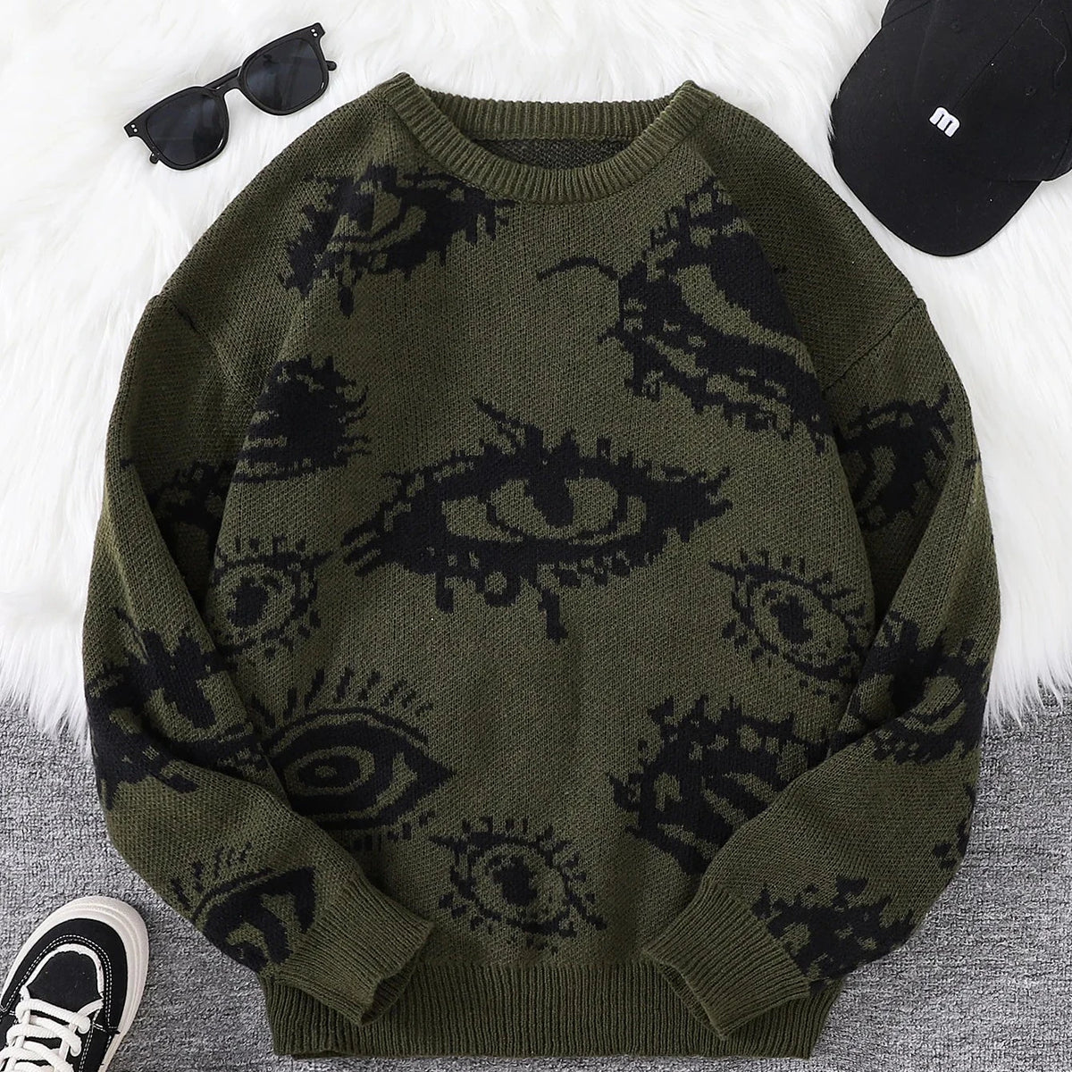 Green sweater with eye pattern, sunglasses, and cap on a textured surface