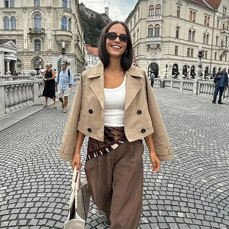 Woman in a beige coat and sunglasses standing on a cobblestone street with historic buildings in the background.