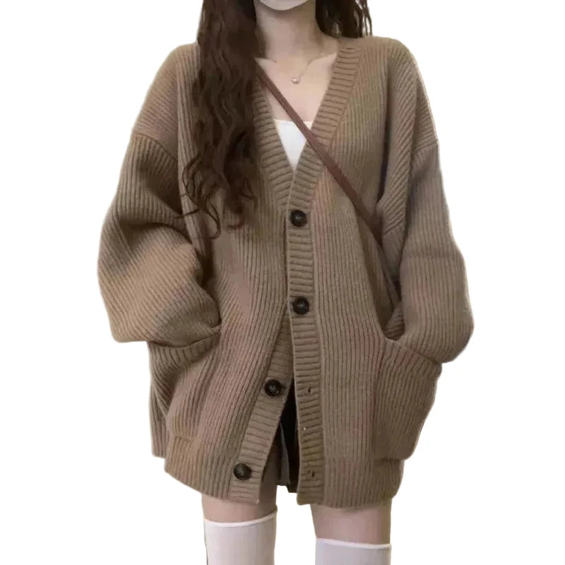 A person wearing an oversized taupe-brown ribbed knit cardigan with large front pockets and dark buttons, layered over a white top and paired with white knee-high socks in an indoor room.