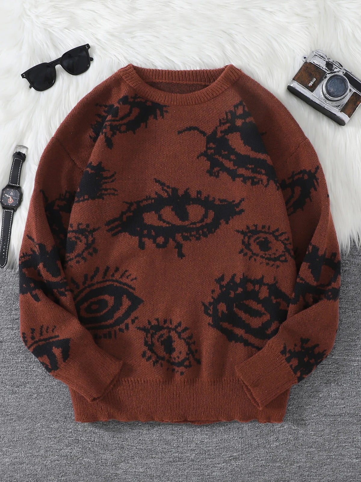 Brown sweater with black eye pattern on a gray surface, surrounded by sunglasses and a camera.