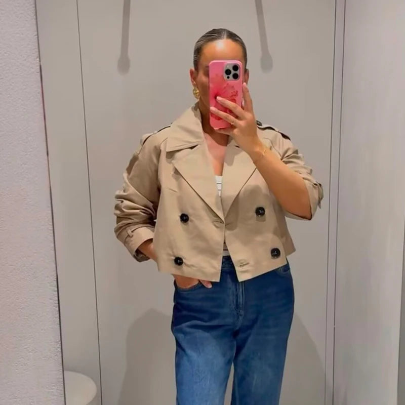 Person wearing a beige trench coat and blue jeans taking a mirror selfie.