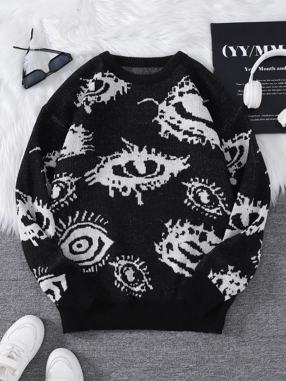 Black sweater with white eye pattern on a gray surface with white shoes and a book in the background.