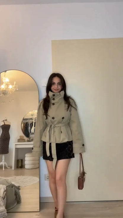 Woman in a beige trench coat standing in a room with a mirror and clothing items.
