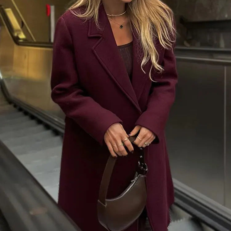 Woman in a purple coat and sunglasses holding a handbag, standing in an indoor setting.