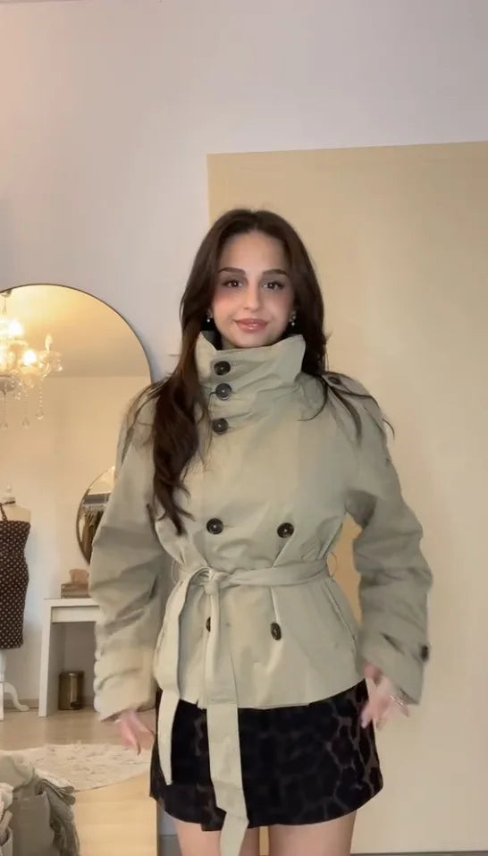 Woman wearing a light-colored trench coat in a room with a mirror and decorative items.