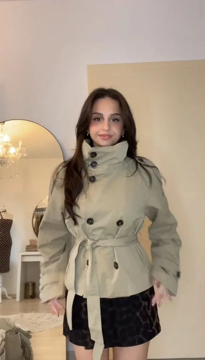 Woman wearing a light-colored trench coat in a room with a mirror and decorative items.