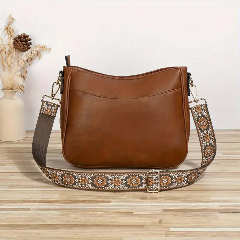 Brown leather handbag with an embroidered strap on a wooden surface.