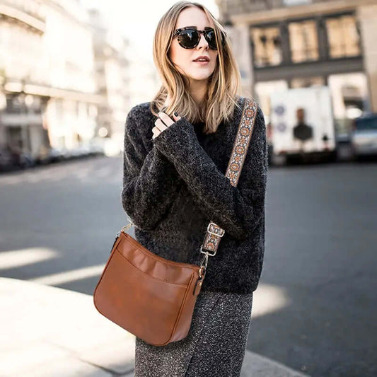 Woman wearing a dark sweater and sunglasses, holding a brown leather bag on a city street.
